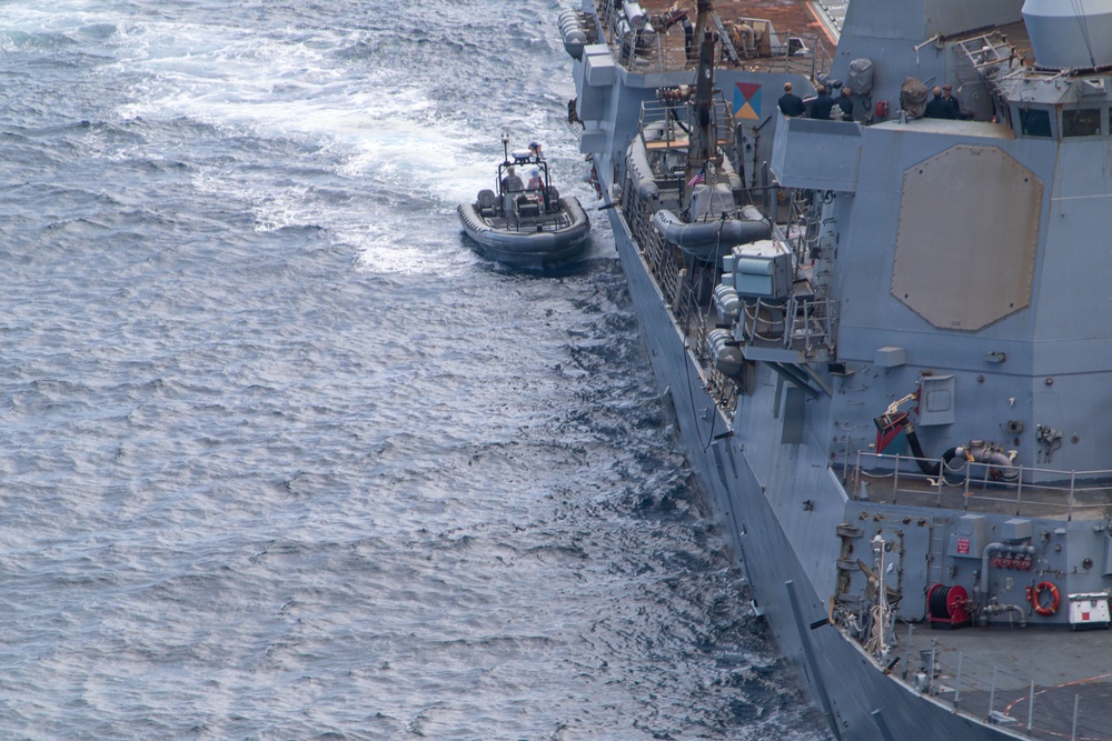 An aerial photograph shows a rigid inflatable boat (RIB) operating alongside a large gray naval warship at sea. The RIB appears to be conducting a small boat operations exercise or transfer mission adjacent to the vessel's port side. The warship displays typical naval destroyer or cruiser characteristics with visible weapons systems, radar equipment, and deck personnel.