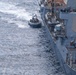 An aerial photograph shows a rigid inflatable boat (RIB) operating alongside a large gray naval warship at sea. The RIB appears to be conducting a small boat operations exercise or transfer mission adjacent to the vessel's port side. The warship displays typical naval destroyer or cruiser characteristics with visible weapons systems, radar equipment, and deck personnel.