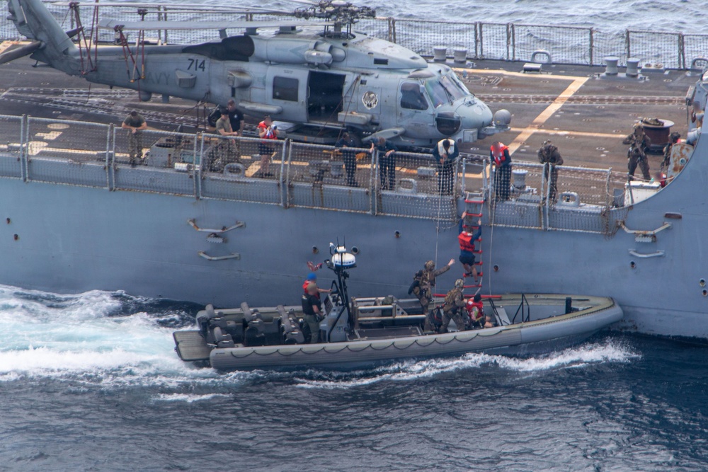 A U.S. Navy rigid-hull inflatable boat (RHIB) is operating alongside a larger naval vessel, with personnel conducting a boarding or transfer operation using a red ladder. A helicopter marked '714' with 'NAVY' visible on its tail is parked on the ship's deck. Personnel in military gear and orange life vests are visible both on the ship's deck and on the RHIB.