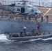 A U.S. Navy rigid-hull inflatable boat (RHIB) is operating alongside a larger naval vessel, with personnel conducting a boarding or transfer operation using a red ladder. A helicopter marked '714' with 'NAVY' visible on its tail is parked on the ship's deck. Personnel in military gear and orange life vests are visible both on the ship's deck and on the RHIB.