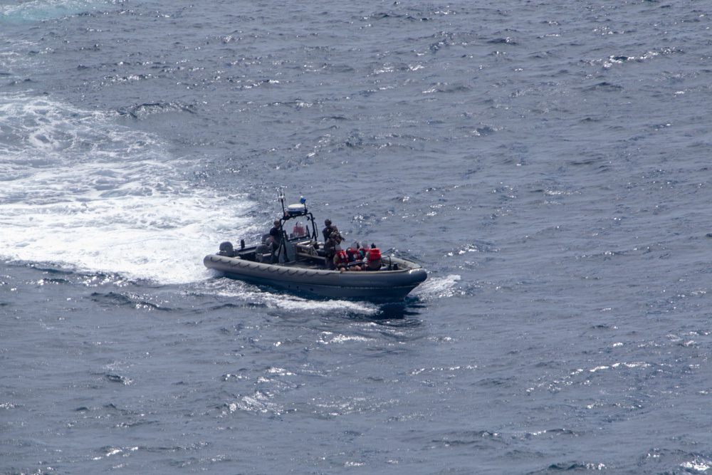 A rigid-hulled inflatable boat (RHIB) with a gray hull is underway at speed on open ocean waters, leaving a visible wake. The vessel carries what appear to be at least two personnel in tactical/military gear along with equipment and red-colored objects aboard. The image is taken from an elevated vantage point, likely from a ship or aircraft.