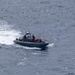 A rigid-hulled inflatable boat (RHIB) with a gray hull is underway at speed on open ocean waters, leaving a visible wake. The vessel carries what appear to be at least two personnel in tactical/military gear along with equipment and red-colored objects aboard. The image is taken from an elevated vantage point, likely from a ship or aircraft.