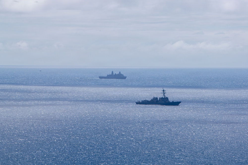 The image shows two naval vessels operating in open ocean under overcast skies. The foreground vessel appears to be a destroyer or guided-missile frigate, while the background vessel is a larger warship, possibly an amphibious transport dock or similar large combatant. Both ships appear to be underway in formation or in proximity during what may be a naval exercise or patrol operation.