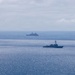 The image shows two naval vessels operating in open ocean under overcast skies. The foreground vessel appears to be a destroyer or guided-missile frigate, while the background vessel is a larger warship, possibly an amphibious transport dock or similar large combatant. Both ships appear to be underway in formation or in proximity during what may be a naval exercise or patrol operation.