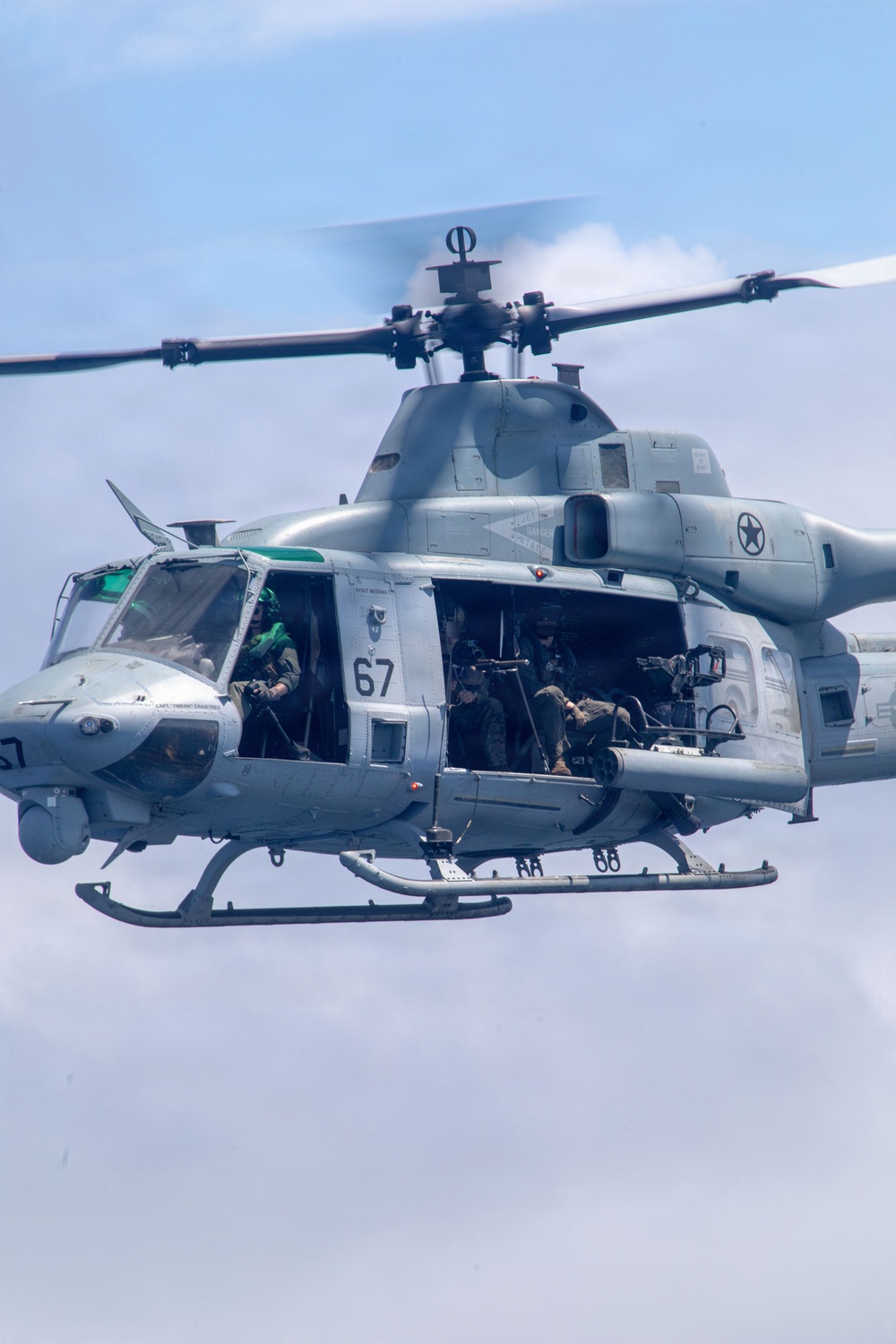 A U.S. military utility helicopter, identifiable as a Bell UH-1Y Venom (bureau number 67), is captured in flight against a partly cloudy sky. The cabin doors are open and armed personnel are visible manning what appears to be a door-mounted weapon system. The aircraft bears U.S. military markings including a star insignia and is painted in standard gray livery.