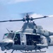 A U.S. military utility helicopter, identifiable as a Bell UH-1Y Venom (bureau number 67), is captured in flight against a partly cloudy sky. The cabin doors are open and armed personnel are visible manning what appears to be a door-mounted weapon system. The aircraft bears U.S. military markings including a star insignia and is painted in standard gray livery.