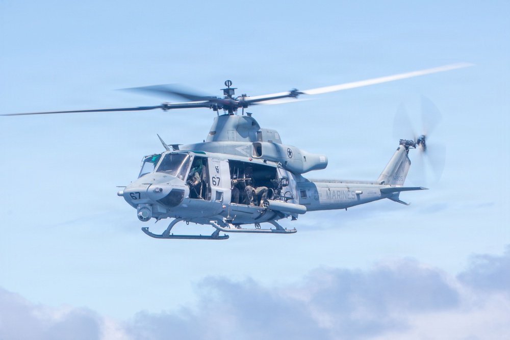 A U.S. Marines military helicopter, marked with the number '67' and 'MARINES' on the fuselage, is captured in flight against a partly cloudy blue sky. The helicopter's side door is open with personnel visible inside, appearing to be operating a door-mounted weapon. The rotor blades show motion blur, indicating active flight.