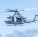 A U.S. Marines military helicopter, marked with the number '67' and 'MARINES' on the fuselage, is captured in flight against a partly cloudy blue sky. The helicopter's side door is open with personnel visible inside, appearing to be operating a door-mounted weapon. The rotor blades show motion blur, indicating active flight.