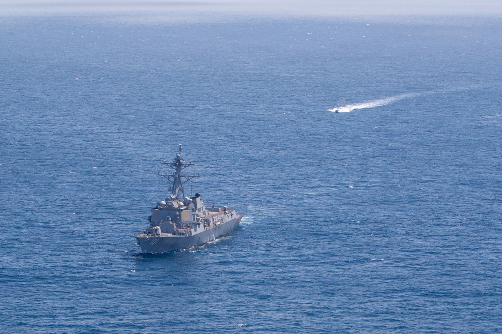An aerial photograph shows a U.S. Navy Arleigh Burke-class guided-missile destroyer underway in open blue ocean waters. A smaller, faster vessel is visible in the upper right portion of the image, leaving a distinct white wake as it moves at high speed. The image appears to capture a naval operation or exercise at sea.
