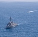 An aerial photograph shows a U.S. Navy Arleigh Burke-class guided-missile destroyer underway in open blue ocean waters. A smaller, faster vessel is visible in the upper right portion of the image, leaving a distinct white wake as it moves at high speed. The image appears to capture a naval operation or exercise at sea.