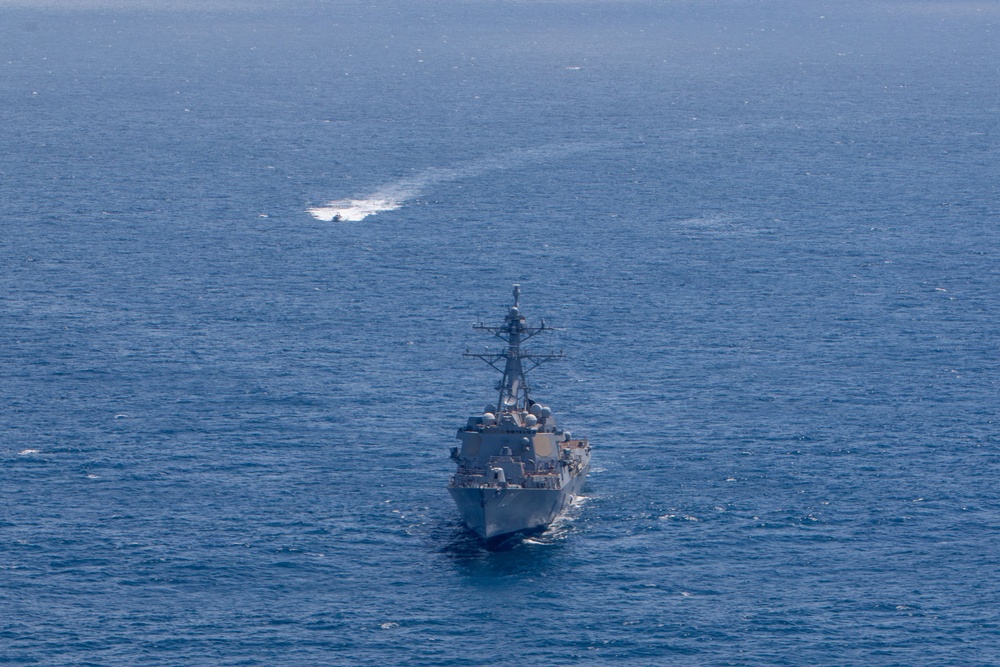 An aerial photograph shows a U.S. Navy warship, consistent with an Arleigh Burke-class guided-missile destroyer, underway in open blue ocean. The ship is photographed from a forward-elevated angle, showing its bow and superstructure clearly. In the background, a second smaller vessel is visible at distance, leaving a white wake trail on the water surface.