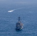 An aerial photograph shows a U.S. Navy warship, consistent with an Arleigh Burke-class guided-missile destroyer, underway in open blue ocean. The ship is photographed from a forward-elevated angle, showing its bow and superstructure clearly. In the background, a second smaller vessel is visible at distance, leaving a white wake trail on the water surface.