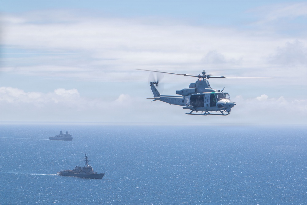 A military helicopter flies over open ocean with two naval vessels visible in the background. The helicopter appears to be a UH-1Y Venom or similar twin-rotor utility helicopter in gray military livery. The scene suggests a naval exercise or patrol operation with coordinated air and sea assets.