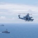 A military helicopter flies over open ocean with two naval vessels visible in the background. The helicopter appears to be a UH-1Y Venom or similar twin-rotor utility helicopter in gray military livery. The scene suggests a naval exercise or patrol operation with coordinated air and sea assets.