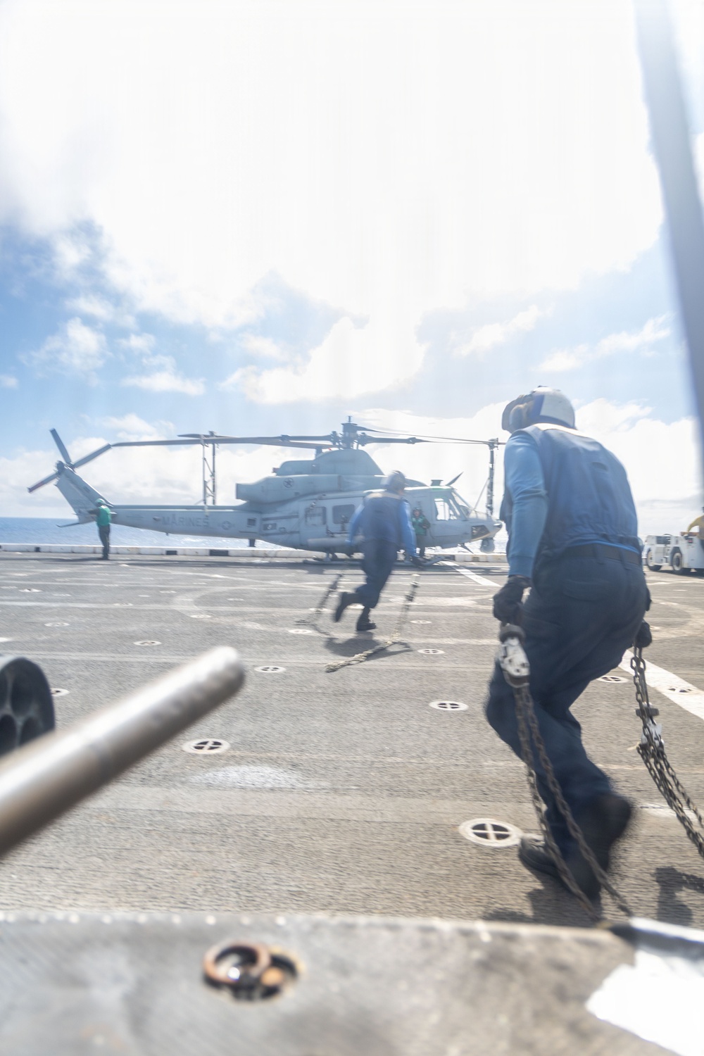 The image shows a flight deck operation aboard a naval vessel, with deck crew members running toward a military helicopter marked 'MARINES'. Personnel wearing blue shirts and protective helmets are carrying chains, likely for securing the aircraft. The scene captures active deck operations under partly cloudy skies over open ocean.