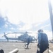 The image shows a flight deck operation aboard a naval vessel, with deck crew members running toward a military helicopter marked 'MARINES'. Personnel wearing blue shirts and protective helmets are carrying chains, likely for securing the aircraft. The scene captures active deck operations under partly cloudy skies over open ocean.
