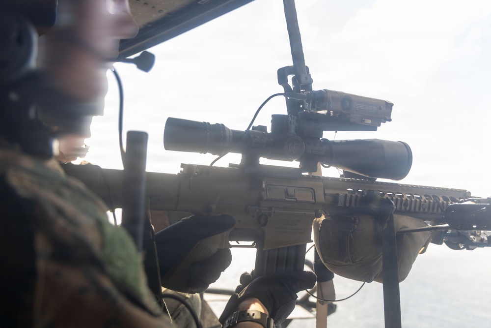A military service member is holding a rifle equipped with a magnified optic scope and additional accessories, appearing to be positioned in or near an aerial platform such as a helicopter. The individual is wearing tactical gear including a helmet with communications headset and camouflage uniform. The background suggests an elevated position with sky and possibly water visible.