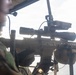 A military service member is holding a rifle equipped with a magnified optic scope and additional accessories, appearing to be positioned in or near an aerial platform such as a helicopter. The individual is wearing tactical gear including a helmet with communications headset and camouflage uniform. The background suggests an elevated position with sky and possibly water visible.