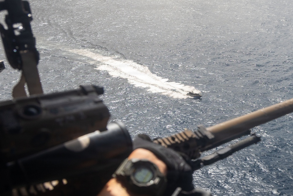 The image is taken from an elevated vantage point, likely a helicopter or ship deck, showing a gunner's position with a mounted weapon system in the foreground. In the background, a small fast-moving boat is visible on open ocean water, leaving a white wake trail. The perspective suggests a military surveillance or overwatch scenario.