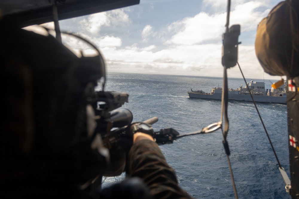 The image is taken from inside a military helicopter, showing an armed personnel aiming a rifle out of the open door toward the ocean. In the background, a naval warship is visible on the water, with what appears to be another helicopter hovering near it. The scene suggests a military exercise or operational scenario involving air and sea assets.