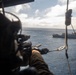 The image is taken from inside a military helicopter, showing an armed personnel aiming a rifle out of the open door toward the ocean. In the background, a naval warship is visible on the water, with what appears to be another helicopter hovering near it. The scene suggests a military exercise or operational scenario involving air and sea assets.