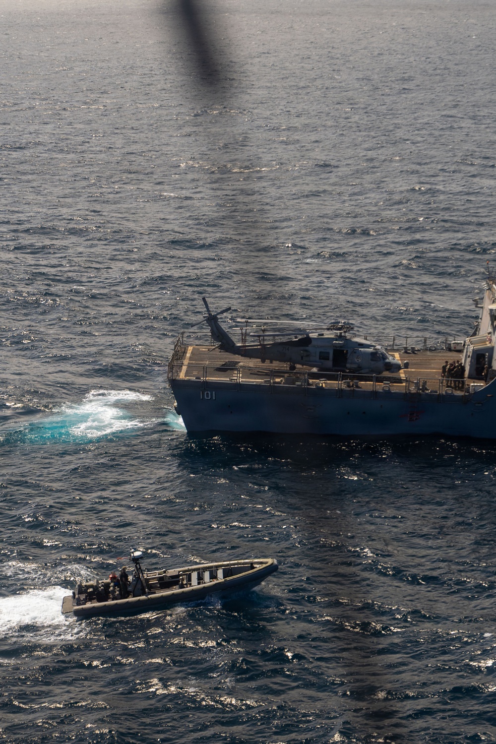 An aerial photograph taken from a helicopter shows a naval vessel numbered '101' operating at sea with a helicopter on its flight deck. A rigid-hull inflatable boat (RHIB) with personnel aboard is operating nearby in the water. The scene appears to depict a maritime military operation, possibly a boarding or interdiction exercise.