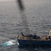 An aerial photograph taken from a helicopter shows a naval vessel numbered '101' operating at sea with a helicopter on its flight deck. A rigid-hull inflatable boat (RHIB) with personnel aboard is operating nearby in the water. The scene appears to depict a maritime military operation, possibly a boarding or interdiction exercise.