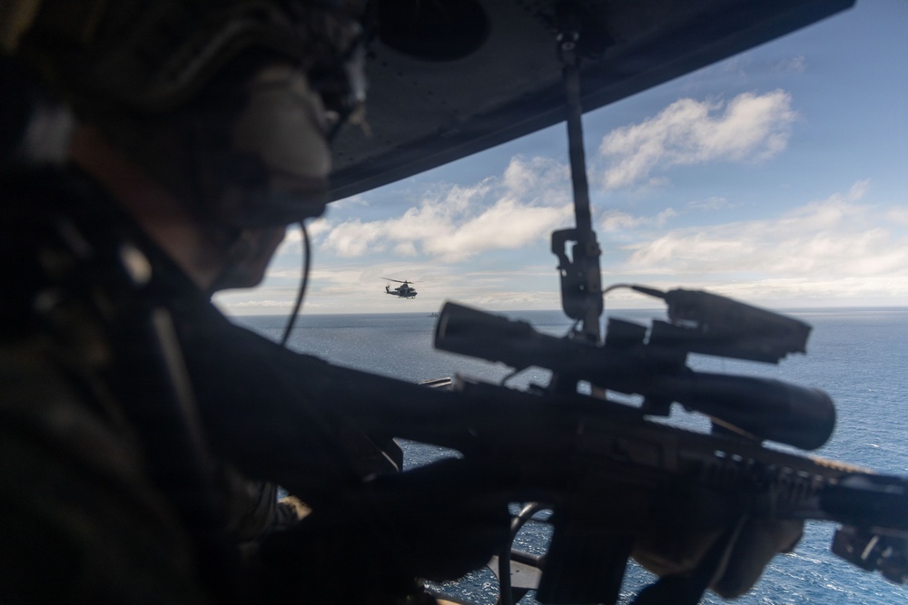 A military personnel is positioned inside a helicopter with a scoped sniper rifle aimed outward through an open door or window. A second helicopter, appearing to be a light attack or utility type, is visible in the mid-distance flying over open ocean. The scene suggests a maritime aerial operation or training exercise.