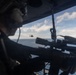 A military personnel is positioned inside a helicopter with a scoped sniper rifle aimed outward through an open door or window. A second helicopter, appearing to be a light attack or utility type, is visible in the mid-distance flying over open ocean. The scene suggests a maritime aerial operation or training exercise.