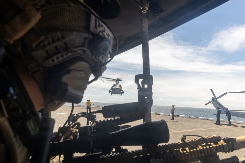 The image is taken from inside a military helicopter looking out onto a naval vessel's flight deck. A gunner in tactical gear with a mounted machine gun is visible in the foreground, while a second helicopter hovers above the flight deck in the background. Flight deck crew in yellow jerseys are visible directing aircraft operations.