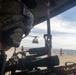 The image is taken from inside a military helicopter looking out onto a naval vessel's flight deck. A gunner in tactical gear with a mounted machine gun is visible in the foreground, while a second helicopter hovers above the flight deck in the background. Flight deck crew in yellow jerseys are visible directing aircraft operations.
