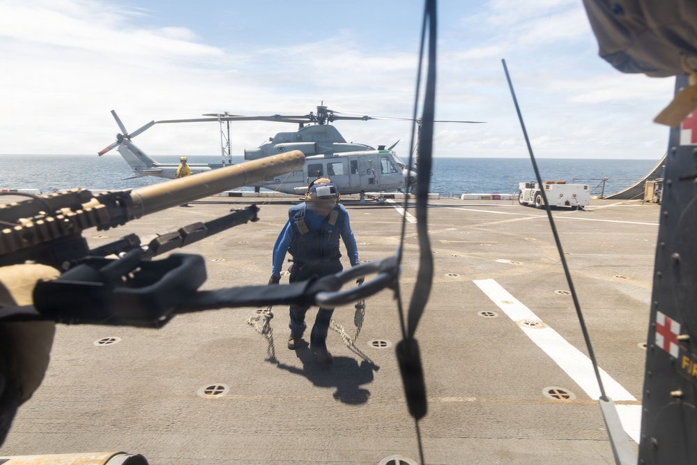 A military naval vessel flight deck is shown with a crew member in blue uniform and hearing protection walking toward the camera while carrying chains or tie-down equipment. A gray military helicopter, consistent with a UH-1Y Venom or similar Bell helicopter, is parked in the background on the deck. The foreground shows what appears to be a deck-mounted weapon system, likely an M2 .50 caliber machine gun.