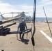 A military naval vessel flight deck is shown with a crew member in blue uniform and hearing protection walking toward the camera while carrying chains or tie-down equipment. A gray military helicopter, consistent with a UH-1Y Venom or similar Bell helicopter, is parked in the background on the deck. The foreground shows what appears to be a deck-mounted weapon system, likely an M2 .50 caliber machine gun.