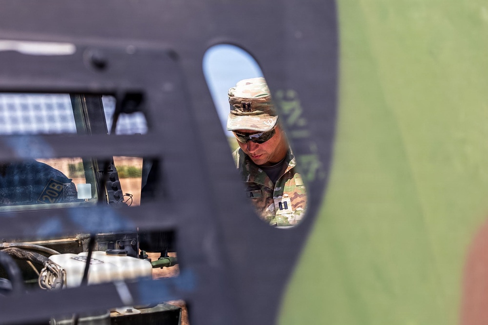 A military personnel member in MultiCam/OCP camouflage uniform and boonie hat is viewed through an oval aperture or opening in a dark metal panel, likely part of a military vehicle or aircraft. The soldier appears to be inspecting or working on equipment, with rank insignia visible on the uniform suggesting a Captain (two silver bars). The composition creates a framed, layered perspective with vehicle components visible in the foreground.