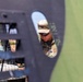A military personnel member in MultiCam/OCP camouflage uniform and boonie hat is viewed through an oval aperture or opening in a dark metal panel, likely part of a military vehicle or aircraft. The soldier appears to be inspecting or working on equipment, with rank insignia visible on the uniform suggesting a Captain (two silver bars). The composition creates a framed, layered perspective with vehicle components visible in the foreground.