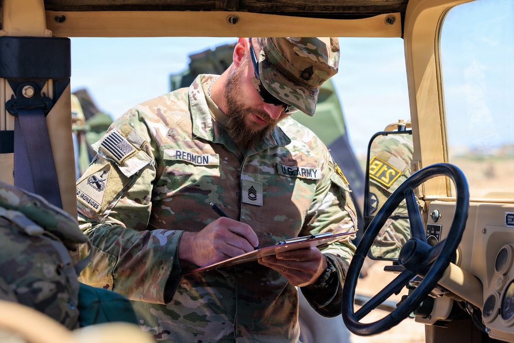 A U.S. Army Staff Sergeant named Redmon, wearing Operational Camouflage Pattern (OCP) uniform, is writing on a clipboard while positioned inside or alongside a military vehicle, likely a HMMWV (Humvee). He is wearing sunglasses and a patrol cap, and his uniform displays a U.S. flag patch, a unit patch reading 'Old Ironsides,' and Staff Sergeant rank insignia. The setting appears to be an outdoor military training or operational environment.