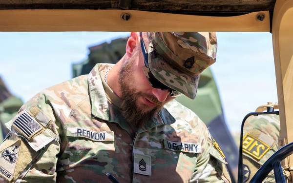 A U.S. Army Staff Sergeant named Redmon, wearing Operational Camouflage Pattern (OCP) uniform, is writing on a clipboard while positioned inside or alongside a military vehicle, likely a HMMWV (Humvee). He is wearing sunglasses and a patrol cap, and his uniform displays a U.S. flag patch, a unit patch reading 'Old Ironsides,' and Staff Sergeant rank insignia. The setting appears to be an outdoor military training or operational environment.