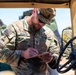 A U.S. Army Staff Sergeant named Redmon, wearing Operational Camouflage Pattern (OCP) uniform, is writing on a clipboard while positioned inside or alongside a military vehicle, likely a HMMWV (Humvee). He is wearing sunglasses and a patrol cap, and his uniform displays a U.S. flag patch, a unit patch reading 'Old Ironsides,' and Staff Sergeant rank insignia. The setting appears to be an outdoor military training or operational environment.