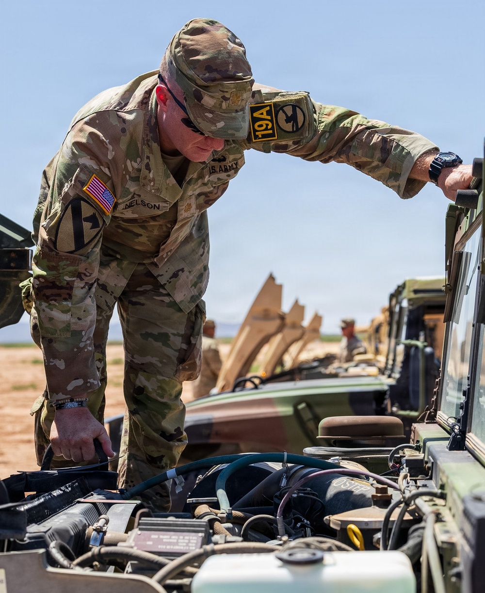 A U.S. Army soldier named Nelson, affiliated with the 1st Cavalry Division and wearing a '19A' branch insignia patch, is inspecting the engine compartment of a military vehicle. The setting appears to be an arid, open training or operational area with a line of military vehicles visible in the background. The soldier is leaning over the open hood, actively examining or adjusting engine components.