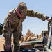 A U.S. Army soldier named Nelson, affiliated with the 1st Cavalry Division and wearing a '19A' branch insignia patch, is inspecting the engine compartment of a military vehicle. The setting appears to be an arid, open training or operational area with a line of military vehicles visible in the background. The soldier is leaning over the open hood, actively examining or adjusting engine components.