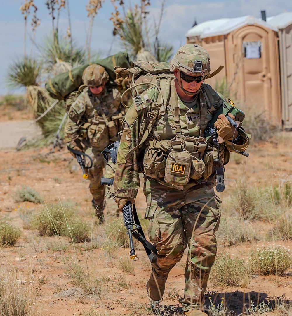 Two U.S. Army soldiers in full combat gear are moving through an arid, desert-like training environment. The lead soldier carries an M4-style rifle and wears a chest rig marked '10A', along with a large rucksack and tactical communications equipment. The setting appears to be a military training exercise, evidenced by the portable toilet in the background and the absence of active combat indicators.
