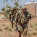Two U.S. Army soldiers in full combat gear are moving through an arid, desert-like training environment. The lead soldier carries an M4-style rifle and wears a chest rig marked '10A', along with a large rucksack and tactical communications equipment. The setting appears to be a military training exercise, evidenced by the portable toilet in the background and the absence of active combat indicators.