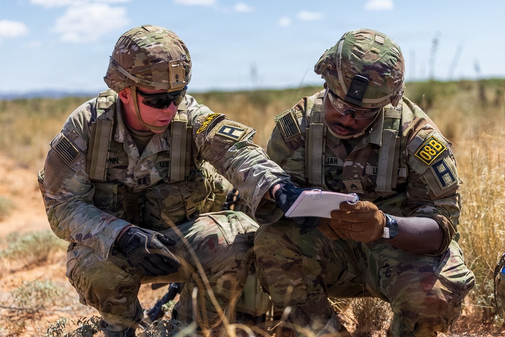 Two U.S. Army soldiers in full combat gear are crouching in an arid, open field while reviewing a document or notepad together. Both wear Operational Camouflage Pattern (OCP) uniforms, tactical vests, helmets, and protective eyewear. The setting appears to be a training exercise or field operation in a dry, scrubland environment.