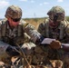 Two U.S. Army soldiers in full combat gear are crouching in an arid, open field while reviewing a document or notepad together. Both wear Operational Camouflage Pattern (OCP) uniforms, tactical vests, helmets, and protective eyewear. The setting appears to be a training exercise or field operation in a dry, scrubland environment.