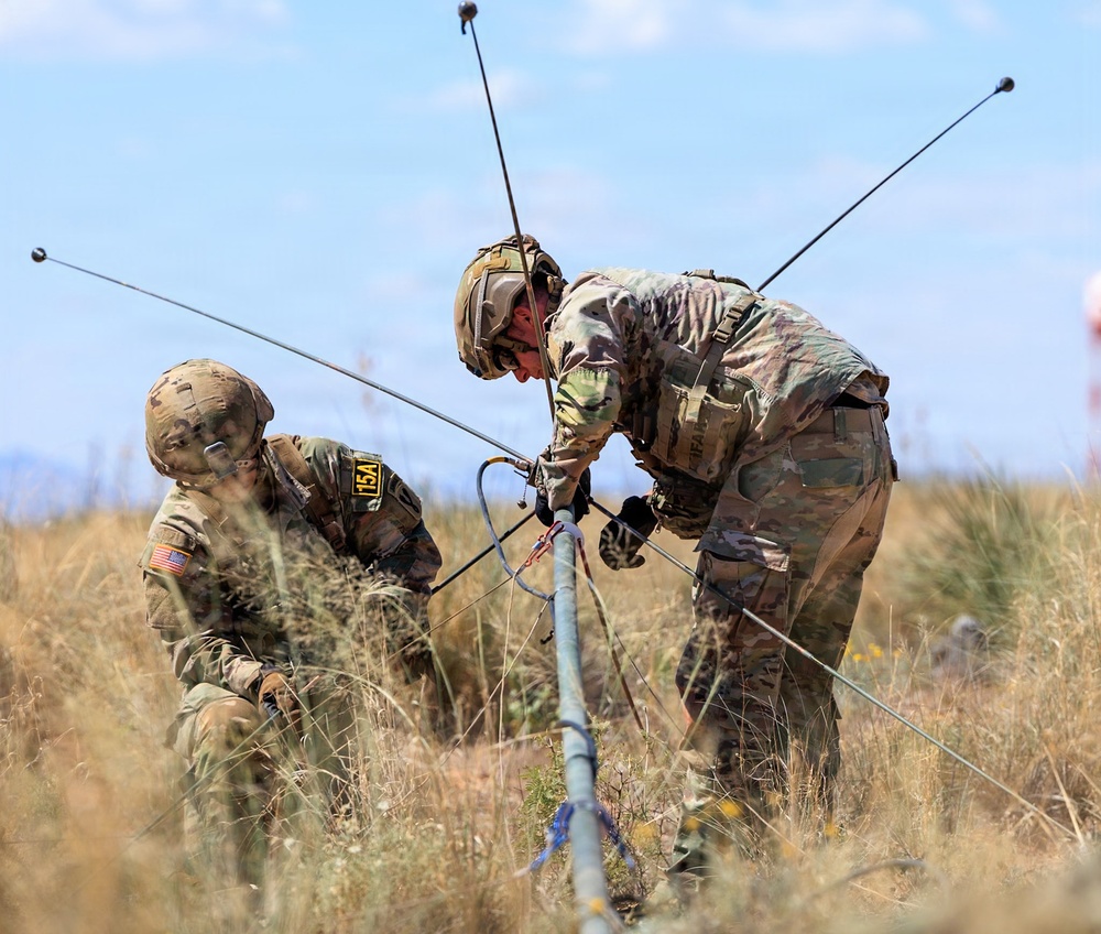 Two U.S. Army soldiers in full combat gear and camouflage uniforms are working together in an open field to assemble or deploy a multi-element antenna system. The soldiers are wearing helmets and tactical vests, with one kneeling and one bending over the equipment. The setting appears to be a dry, grassy terrain under a partly cloudy sky, consistent with a field training exercise.