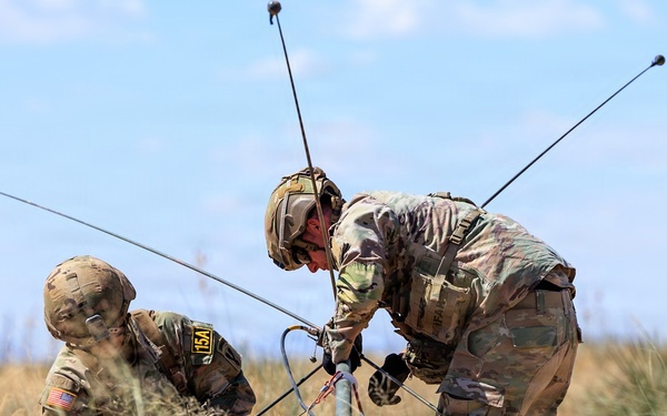 Two U.S. Army soldiers in full combat gear and camouflage uniforms are working together in an open field to assemble or deploy a multi-element antenna system. The soldiers are wearing helmets and tactical vests, with one kneeling and one bending over the equipment. The setting appears to be a dry, grassy terrain under a partly cloudy sky, consistent with a field training exercise.