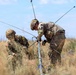 Two U.S. Army soldiers in full combat gear and camouflage uniforms are working together in an open field to assemble or deploy a multi-element antenna system. The soldiers are wearing helmets and tactical vests, with one kneeling and one bending over the equipment. The setting appears to be a dry, grassy terrain under a partly cloudy sky, consistent with a field training exercise.