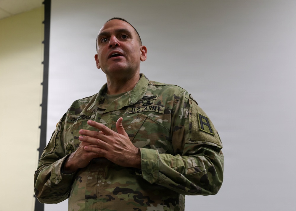 A U.S. Army soldier in Operational Camouflage Pattern (OCP) uniform is speaking or briefing, with hands clasped at chest level in an expressive gesture. The name tape reads 'DIAZ-PENA' and the uniform displays a 'U.S. ARMY' tape, a shoulder patch, and various badges. The setting appears to be an indoor facility such as a classroom or briefing room.