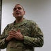 A U.S. Army soldier in Operational Camouflage Pattern (OCP) uniform is speaking or briefing, with hands clasped at chest level in an expressive gesture. The name tape reads 'DIAZ-PENA' and the uniform displays a 'U.S. ARMY' tape, a shoulder patch, and various badges. The setting appears to be an indoor facility such as a classroom or briefing room.