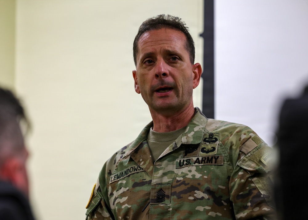 A U.S. Army soldier wearing Operational Camouflage Pattern (OCP) uniform is speaking, likely addressing a group. The name tape reads 'LEWANDOWS' (partially visible, likely Lewandowski) and rank insignia on the collar indicates a senior NCO rank. A Master Parachutist badge is visible on the chest.